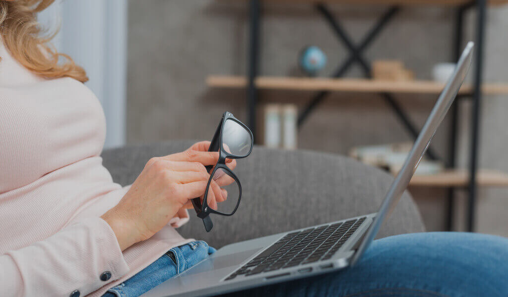 Woman holding eyeglasses in hand sitting on sofa with an open laptop on her lap and writing SRS